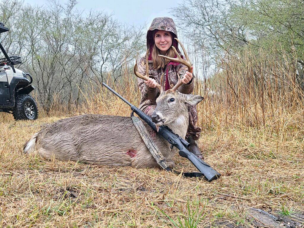 Jessica holding up 8-point buck with rifle on the ground